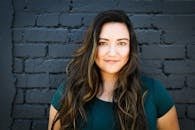 A captivating portrait of a woman smiling confidently in front of a brick wall, showcasing her long brunette hair and vibrant eyes.